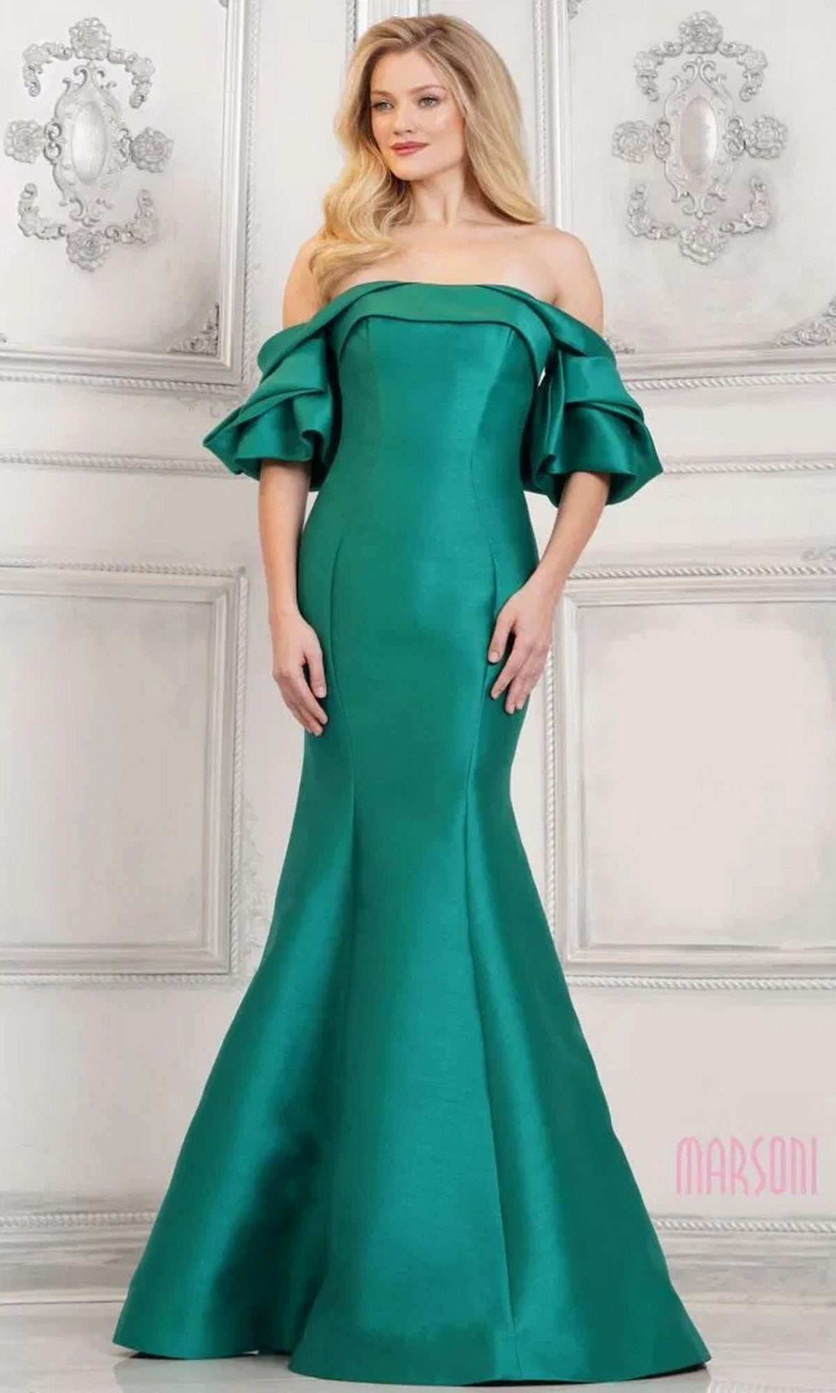 A woman is wearing the Marsoni MV1290 long formal dress in green dupioni silk with ruffled sleeves and a mermaid silhouette, standing before an ornate light-colored wall.